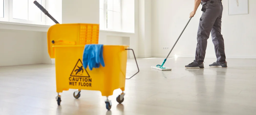 A person mops a light-colored floor in a bright room. In the foreground, there is a yellow mop bucket on wheels with a blue cloth and a "Caution Wet Floor" sign printed on it.