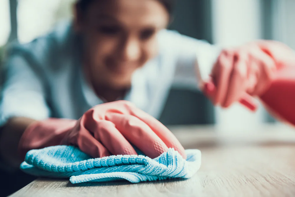 A person wearing pink rubber gloves is smiling while cleaning a wooden surface with a blue microfiber cloth, focusing closely on their hands and the cloth.