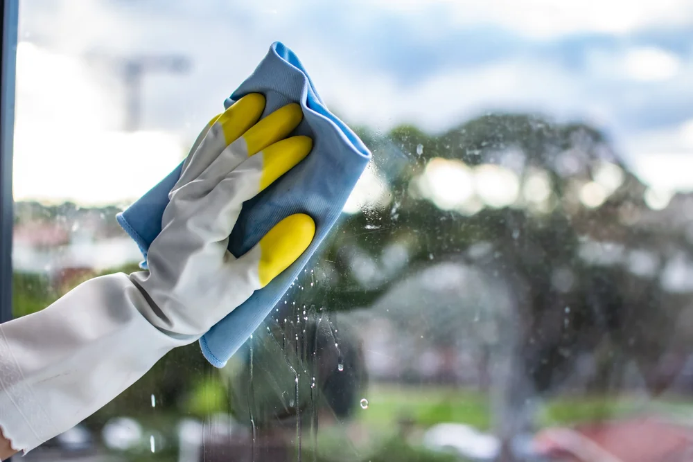 A person wearing yellow and white rubber gloves is cleaning a window with a blue microfiber cloth. The window has water spots, and an outdoor scene with trees is visible in the blurred background.