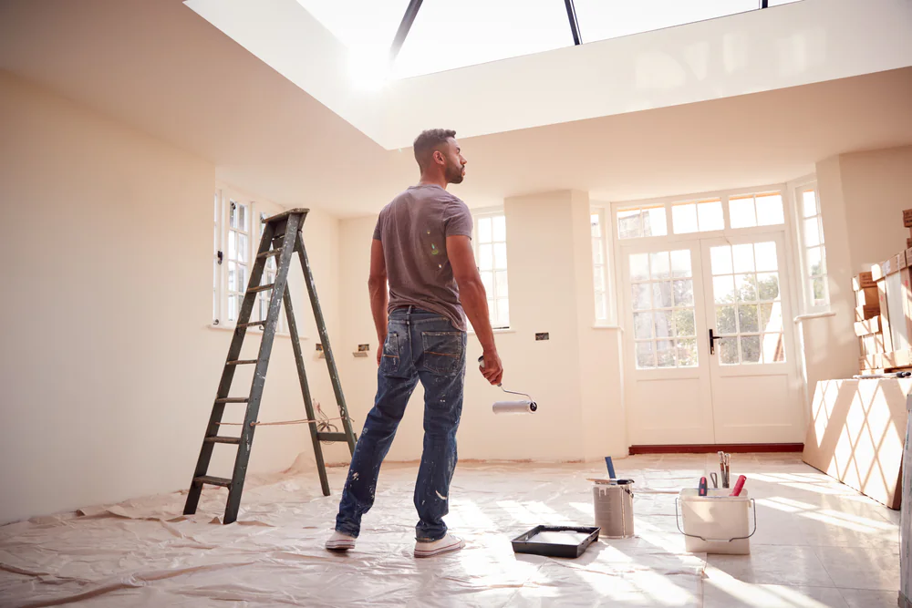 A man holding a rolled-up paper stands in a bright room under renovation, with a ladder, paint supplies, and protective sheets covering the floor. Sunlight streams in through large windows and a skylight.
