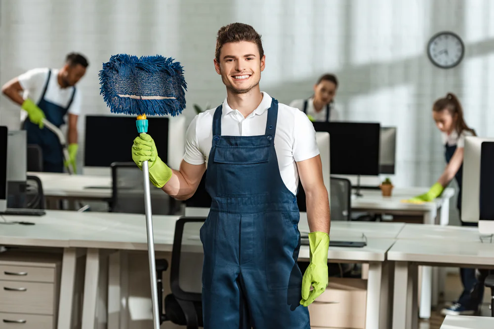 A smiling young man in overalls and rubber gloves holds a mop in an office, with three other people cleaning desks and computers in the background.