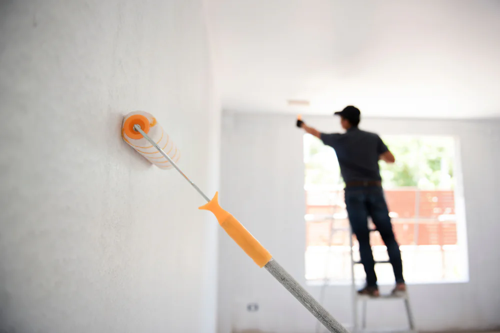 A close-up of a paint roller applying white paint to a wall, with a person standing on a ladder and working near a window in the blurred background.