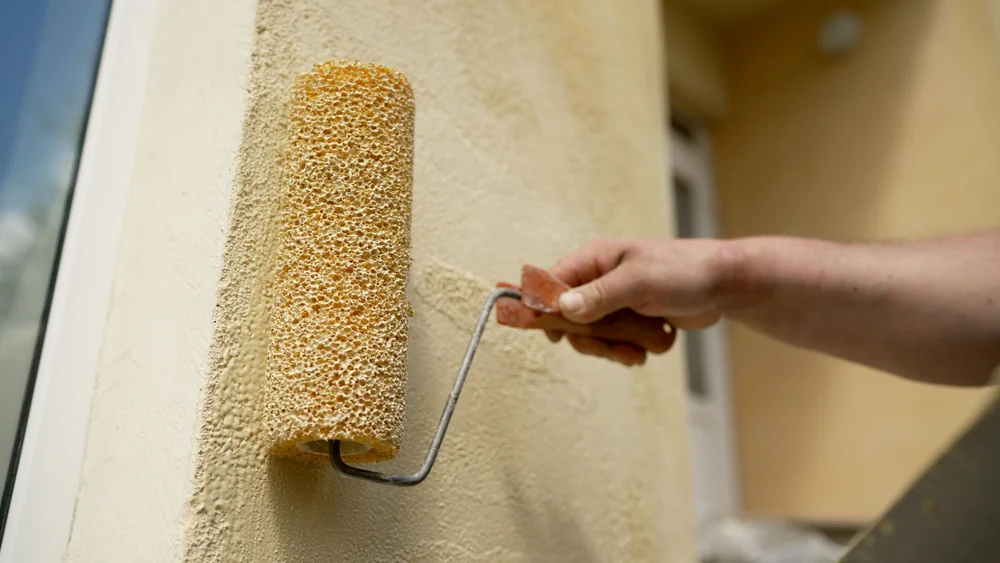 A close-up of a hand holding a paint roller, applying a light beige color to a textured exterior wall. The background shows part of a doorway and blurred building details.