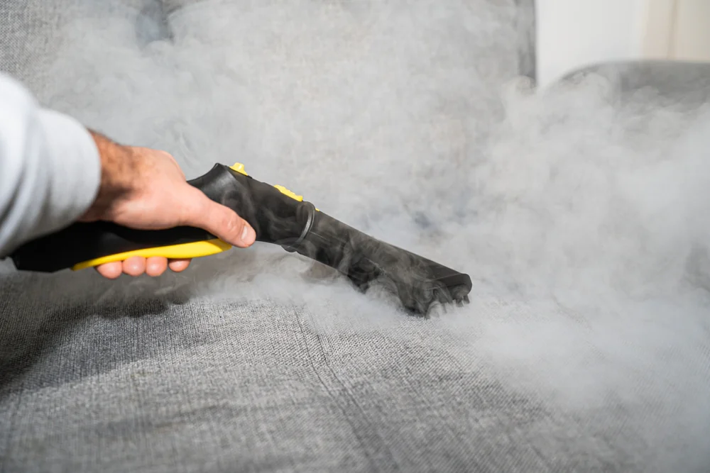 A person uses a handheld steam cleaner on a gray fabric couch, releasing visible steam to clean and disinfect the upholstery.